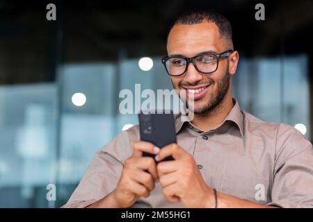 Close-up of smiling African-American businessman using phone typing messages and browsing internet pages, man in shirt and glasses smiling happily inside office holding smartphone. Stock Photo