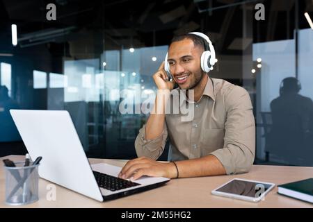 Young hispanic businessman in shirt working inside office using laptop at work, man with headphones listening to music and audio podcasts at work. Stock Photo