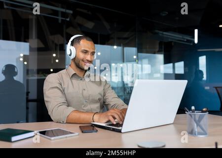 Young hispanic businessman in shirt working inside office using laptop at work, man with headphones listening to music and audio podcasts at work. Stock Photo