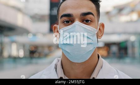 Hispanic Indian Ethnic doctor man medic in medical face protective mask uniform male therapist surgeon dentist nurse looking to camera indoors posing Stock Photo
