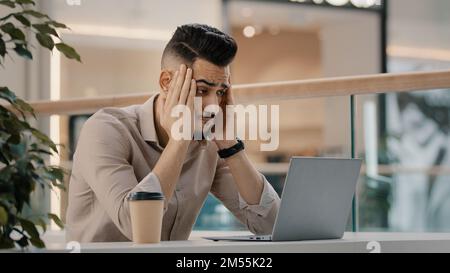 Sad shocked stressed ethnic CEO Indian man worker manager freelancer entrepreneur manager in cafe has problem with laptop app reading bad news worried Stock Photo