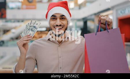 Indian happy Hispanic guy man in Santa X-mas Christmas hat in shopping mall looking camera showing holding money cash dollars and gift bags male Stock Photo