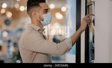 Hispanic Indian passenger male buyer man in medical mask in airport cafe touching self-service machine in buying ticket make online order scroll touch Stock Photo