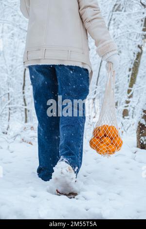 In the snow, a woman holds a wicker bag filled with tangerines next to here feet as she walks Stock Photo
