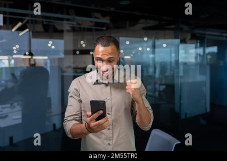 Businessman is happy to celebrate victory and success, Hispanic man inside the office holds a smartphone in his hands received a joyful message news of triumph holds his hand up. Stock Photo