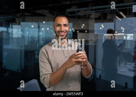Portrait young hispanic man inside office man in a casual shirt is smiling and looking at the camera, a businessman is holding a smartphone in his hands, typing a message on the phone, making a call. Stock Photo