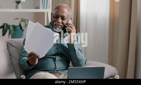 African biracial bald old man with gray beard senior mature businessman busy employee using laptop talking cell phone in multitasking discussing Stock Photo