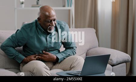 Older african american male freelancer mature pensioner sitting at home office on couch using laptop feel acute stomach pain discomfort indigestion Stock Photo