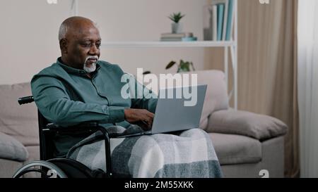 Pensive though in deep thinking. old 60s American businessman sitting at home in wheelchair online working typing on laptop writing text novel report Stock Photo
