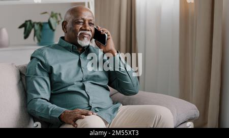 African bald middle-aged man senior mature businessman sitting on sofa at home in office talking phone chatting mobile conversation making order with Stock Photo