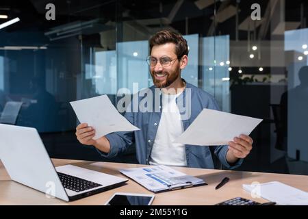 Satisfied with the result financier businessman holds in his hands and examines contracts and reports smiling and happy, man in a casual shirt works inside the office using a laptop in paperwork. Stock Photo