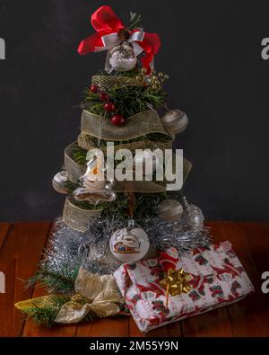 A present is placed under a synthetic Christmas tree decorated with balls of various colors and a red bow on top Stock Photo