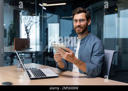 Portrait of a businessman in a modern office, a man uses a tablet computer, sits at a desk, smiles and looks at the camera in a casual shirt and glasses. Stock Photo
