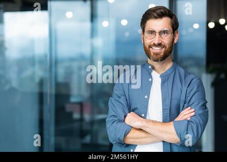 Portrait of successful businessman inside modern office, mature man with beard and glasses smiling and looking at camera, boss in casual clothes shirt near window with crossed arms. Stock Photo