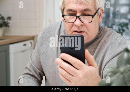 Hoary old man looking shouting to web camera, holding phone talking with clients online. Senior male is serious chatting with partners via video call. Stock Photo