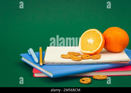 Notebooks and orange on green background, school snack concept. Stock Photo