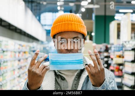 Young African-American man in stylish knitted hat puts on blue disposable mask standing in supermarket department close view Stock Photo