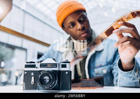 Concentrated African-American man photographer in denim jacket checks vintage film at table focus on old camera close view Stock Photo