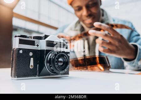 Positive African-American guy photographer in denim jacket checks vintage film sitting at table focus on old camera closeup Stock Photo