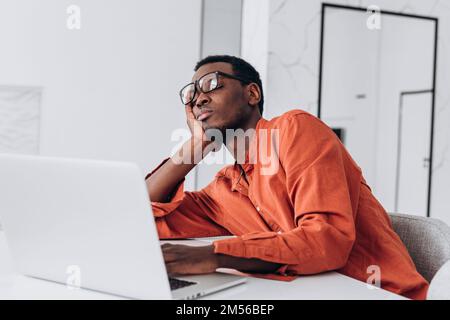 Tired young African-American man with glasses sleeps leaning on hand near laptop at white table in light room close view Stock Photo