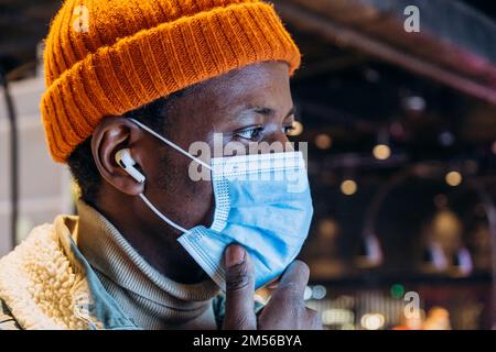 Attractive African-American guy in hat with protective mask and wireless earphones touches chin on blurred background closeup Stock Photo