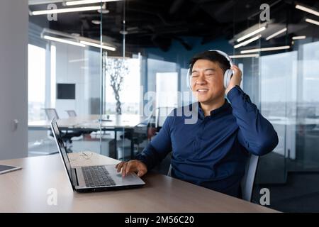 Young man Asian businessman, freelancer sits in the office at the table, works on a laptop, listens to music in white headphones, closed his eyes, enjoys, rests. Stock Photo