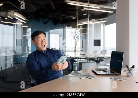 A happy young Asian man, an office worker, an entrepreneur holds cash money in his hands, received a salary, a bonus. He is sitting at a table in the office, rejoices, shouts with joy. Stock Photo