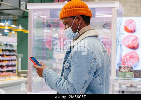 African-American man in stylish clothes with protective mask uses mobile phone in supermarket meat department side view Stock Photo