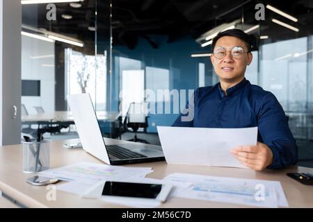 Portrait of a young Asian architect, designer, engineer. Sitting at the desk in the office, holding papers. Works with documents, projects, plans. He looks at the camera, smiles. Stock Photo