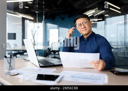 Serious young Asian architect, designer, engineer. Sitting at the desk in the office, holding documents and a pencil. The problem is working with projects, plans. Looking worriedly at the camera. Stock Photo