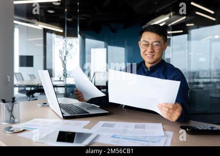 A young Asian architect, designer, engineer. Sitting at the desk in the office, holding papers. Works with projects, plans. Studying documents, deal. Stock Photo