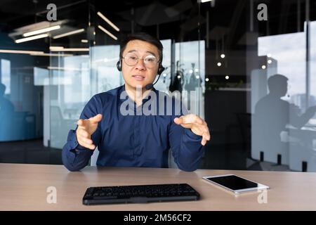 A young Asian man sits in the office at a desk in a headset, looks and talks to the camera. Explains, advises, conducts training, online meeting. Stock Photo