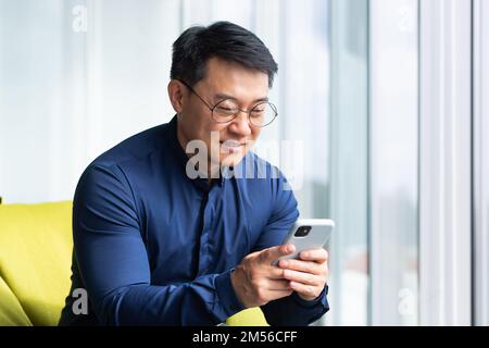 A young man, an Asian student, sits on an ottoman by the window in an office, co-working space, campus. He holds the phone in his hands, dials a message, reads the news, checks the mail, smiles. Stock Photo