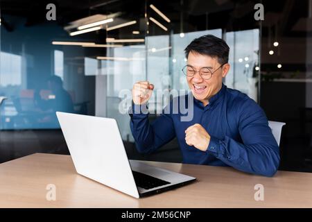 A young male Asian student received good news, mail, passed the exam, entered the university, received a scholarship. He sits in the office, looks at the monitor, rejoices, shows yes with his hand Stock Photo