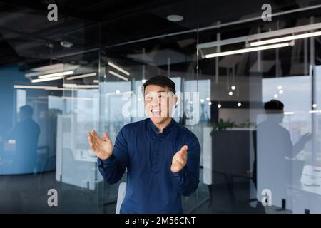 Young man Asian businessman, employee dancing in the office, celebrating victory, successful deal, monetary reward, promotion. He smiles, claps his hands. Stock Photo