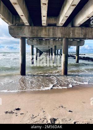 A vertical shot of sea waves hitting the shore of a beach seen from under a wooden pier Stock Photo
