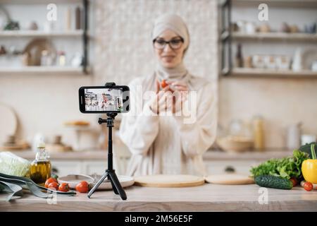 Focus on smartphone, pretty muslim female blogger in casual wear and hijab recording online video on mobile phone, show cooking process of salad, standing at light kitchen at home. Stock Photo