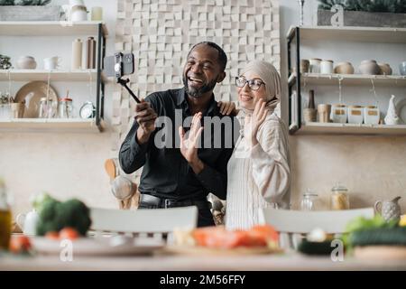 Multiethnic family having video call using selfie stick while providing cooking online class healthy vegan food Happy young couple blogger showing to followers how prepare dinner with fresh vegetable Stock Photo