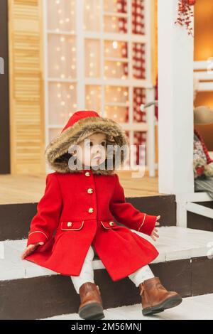 charming baby in a red coat with a hood sits on the porch Stock Photo