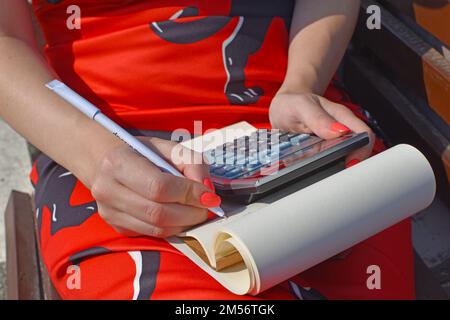A closeup shot of a woman sitting on bench and writing on notebook with using calculator Stock Photo