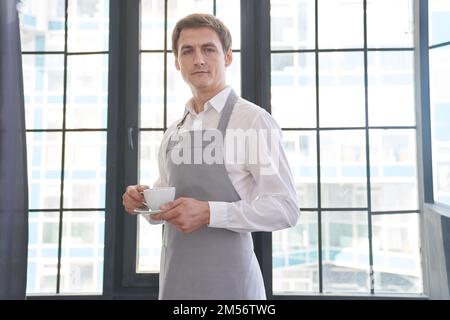 Portrait of a successful young man working as a professional barista, smiling at the camera and holding a coffee drink for a client. Positive male waiter. High quality photo Stock Photo