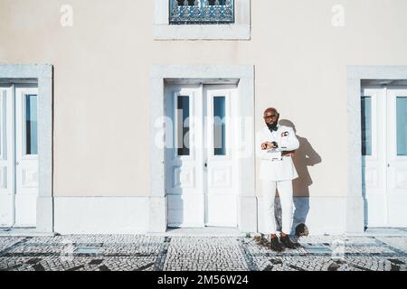 An African-American, well-groomed, bearded male wearing a white suit leaning against the pastel wall of a classic building with tall doors. The busine Stock Photo