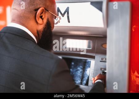 A closeup of a hairless, African American businessman, wearing glasses and a tailored tuxedo with a square pattern, inserting a credit card to withdra Stock Photo