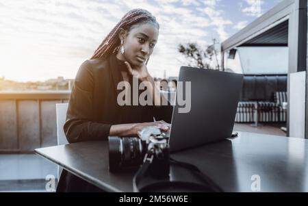 A resplendent young African woman working as a freelancer while sitting on a rooftop cafe, on top of the table both her laptop and camera, with her he Stock Photo