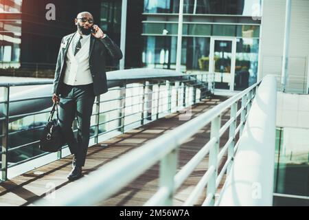 A white-collar worker carries a weekender while having a phone call when leaving the office. A hairless black man wearing sunglasses, a suit, a tie, a Stock Photo