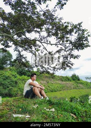 young latin man sitting on top of a mountain looking at the beautiful green colombian landscape, man sitting under a tree Stock Photo