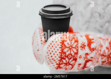 Woman in warm wool clothes drinking hot coffee. Female hands in two-tone winter mittens holding black paper cup. Close up photo with copy space Stock Photo