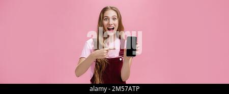 Excited and thrilled young teenage girl in overalls and t-shirt smiling amazed full of happiness pointing at smartphone screen staring enthusiastic at Stock Photo