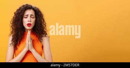 Nervous and concerned woman with curly hairstyle feeling hopeful praying with closed eyes and frowned eyebrows holding hands in pray near chest Stock Photo