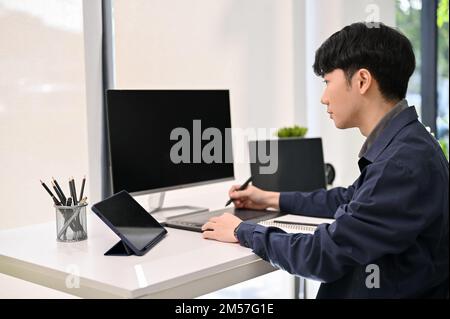Professional and smart young Asian male website developer or programmer focusing on his project, using graphic tablet at his desk. Stock Photo